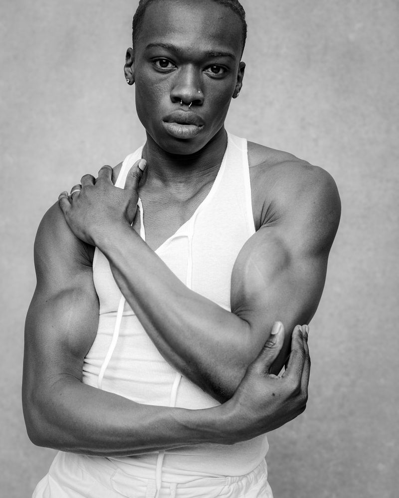 Portrait of a Black man with a nose ring wearing a white tank top against a textured grey background.