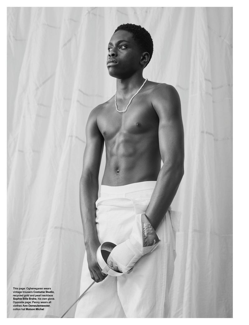 Shirtless young black man holding a fencing foil wearing white trousers against draped fabric background