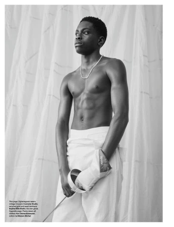 Shirtless young black man holding a fencing foil wearing white trousers against draped fabric background
