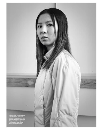 Monochrome portrait of a young woman wearing a padded jacket against an interior wall with wood paneling.