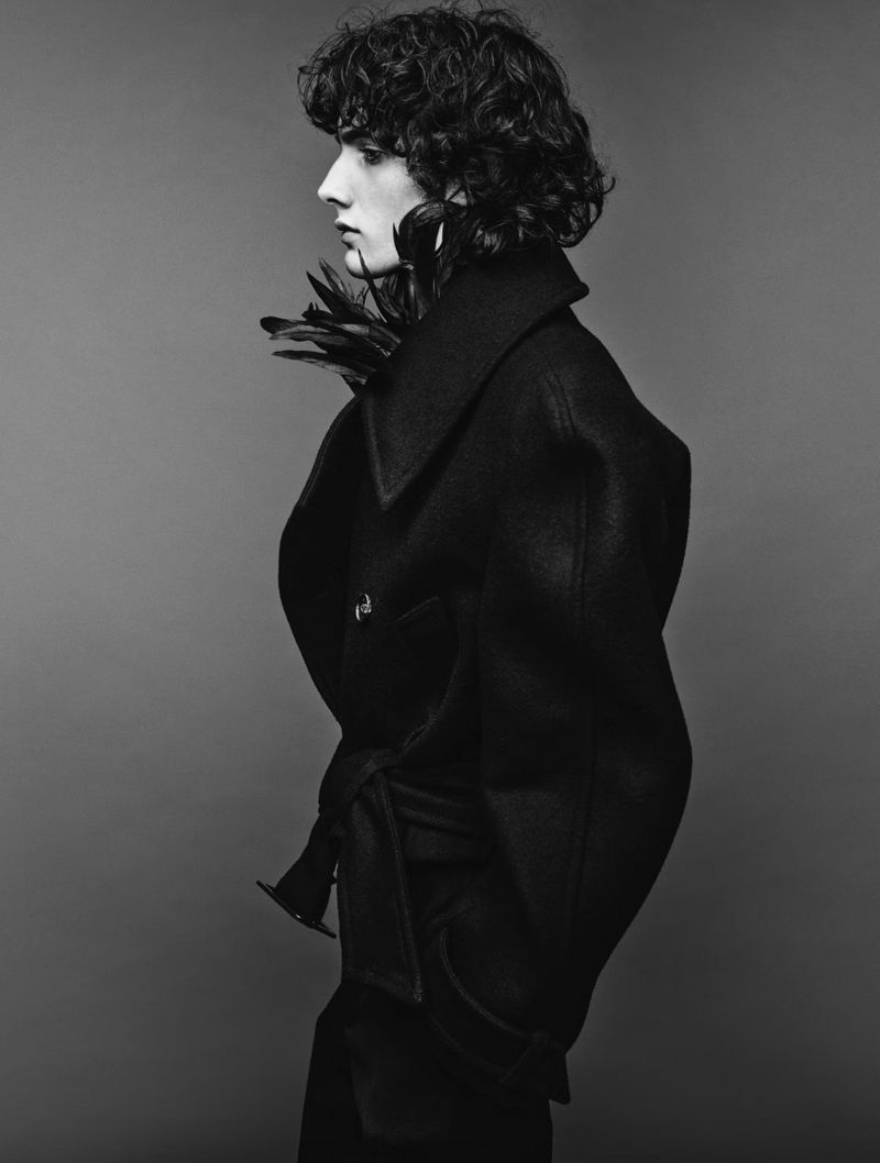 Monochrome studio fashion portrait of a person with curly hair wearing a dark coat with feather collar details against a grey backdrop