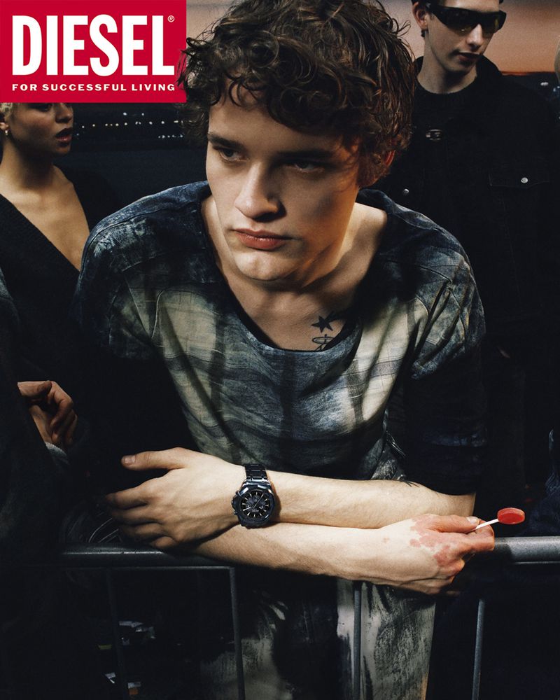 A young man leaning against a railing wearing a patterned top with a black wristwatch, part of a Diesel SS26 campaign image.