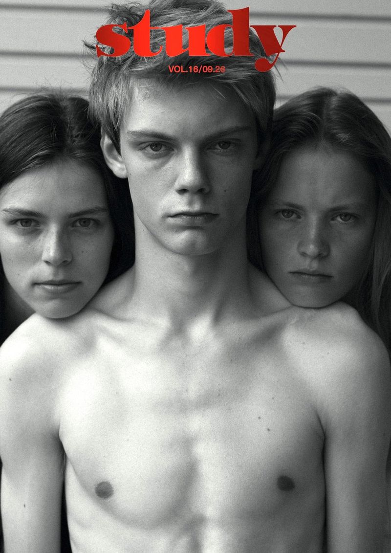 Black and white portrait of a young shirtless man flanked by two women against an exterior wall