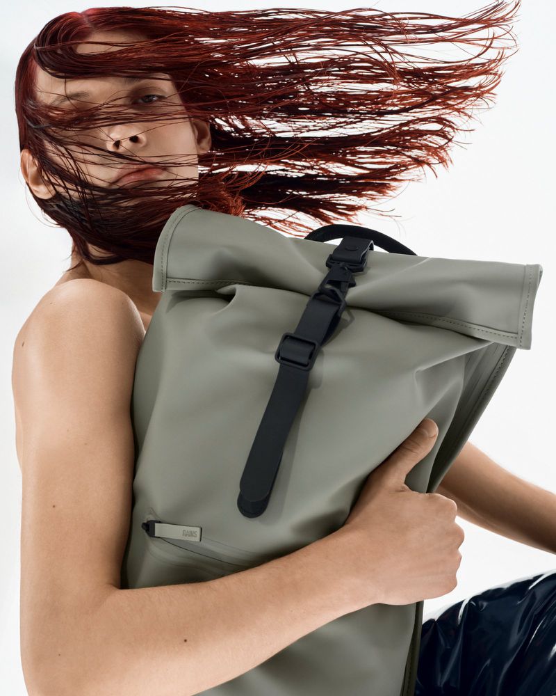 A person poses indoors with a gray roll-top backpack. The person has red hair blowing in the wind, and the backpack is held closely.