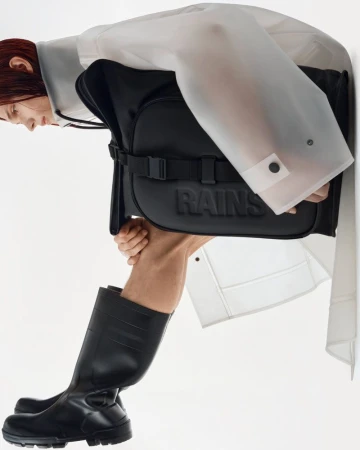Eye-level portrait shot of a person posing in black boots and a transparent jacket holding a black RAINS backpack against a white background.