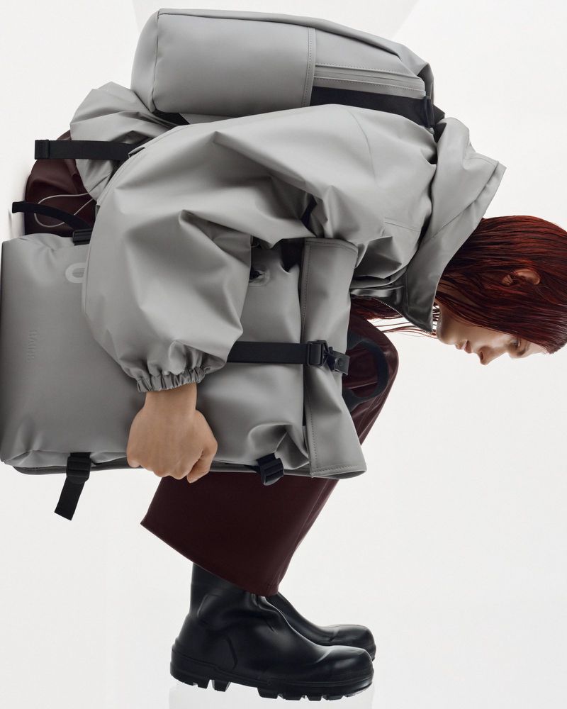 Adult female with auburn hair, wearing a grey coated backpack and black boots, bending over in a brightly lit, stark white studio.