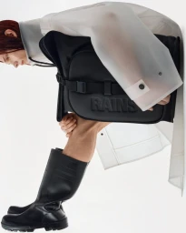 Model wearing a translucent waterproof jacket, tall black boots, and holding a black embossed Rains bag against a white backdrop.