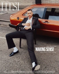 Man wearing a black suit and white socks sitting in a chair beside an orange vintage car on a street