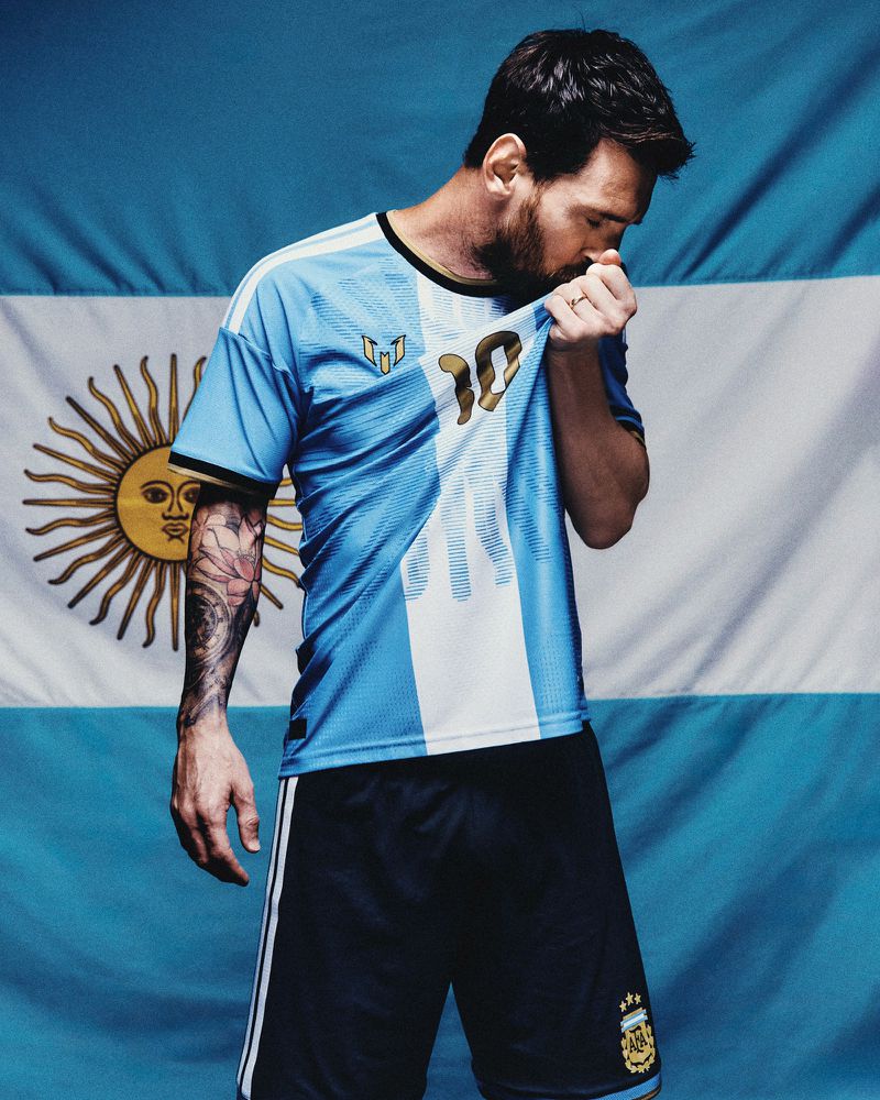 Lionel Messi in an Argentina football jersey pulling his shirt collar against an Argentine flag backdrop.