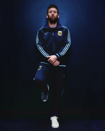 Lionel Messi wearing a navy blue Adidas tracksuit leaning against a dark blue background