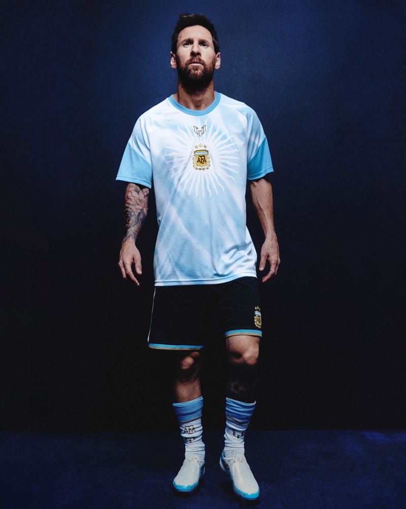 Lionel Messi wearing a light blue and white jersey and dark shorts standing against a dark blue seamless studio background.