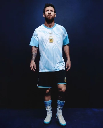 Lionel Messi wearing a light blue and white jersey and dark shorts standing against a dark blue seamless studio background.