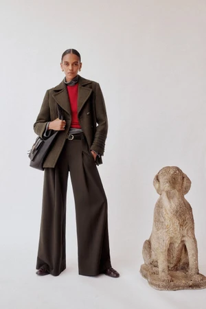 Model standing in an olive green peacoat and wide trousers next to a stone dog statue against a plain backdrop.