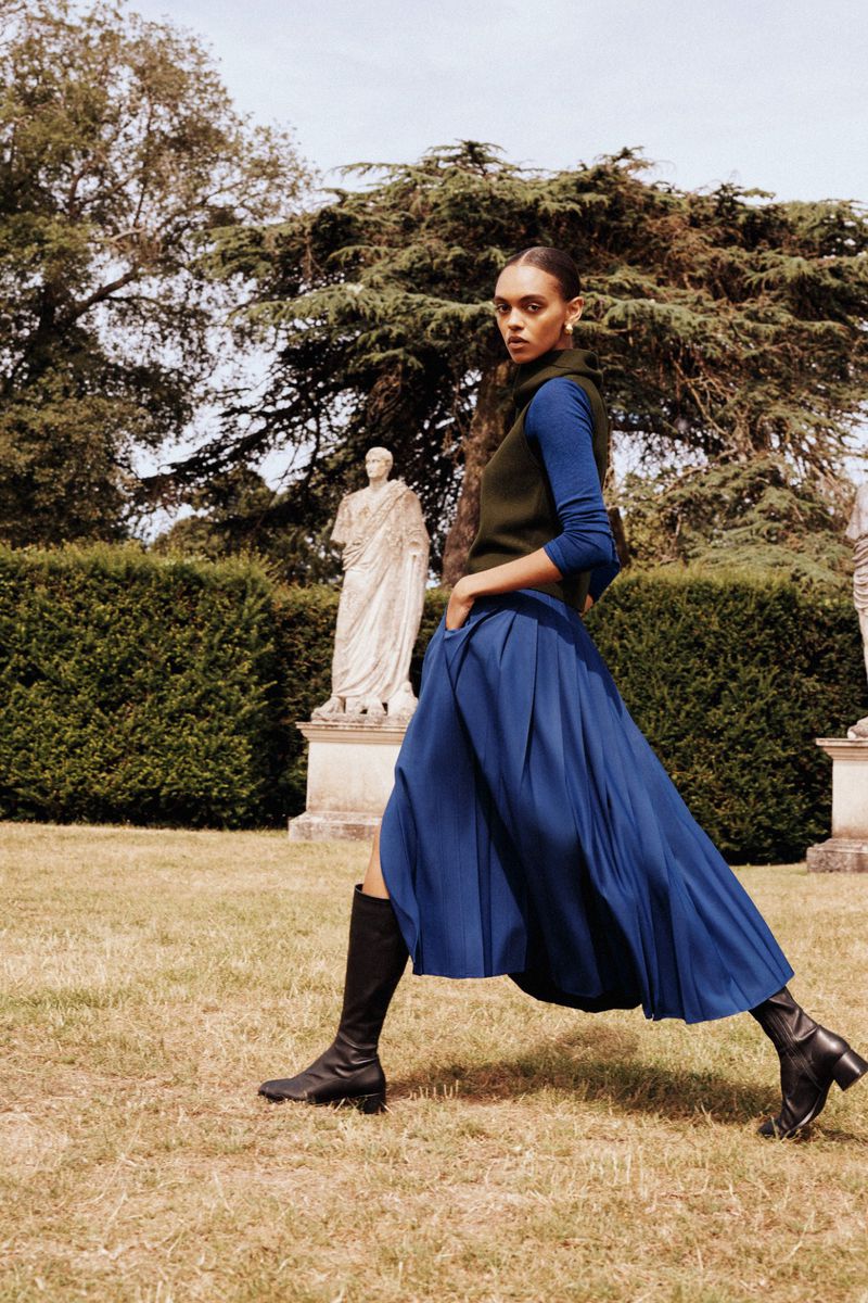 Model wearing a blue pleated skirt and dark boots outdoors on a grassy lawn with classical statues and trees in the background.