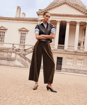 Model standing with arms crossed wearing a leather vest over a button shirt and wide-leg trousers outside a grand building.