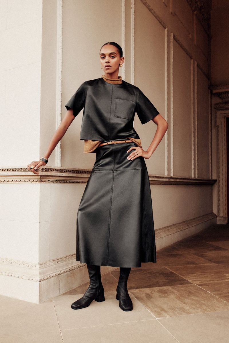 Model wearing a dark leather short-sleeve top and matching midi skirt standing in an ornate hallway