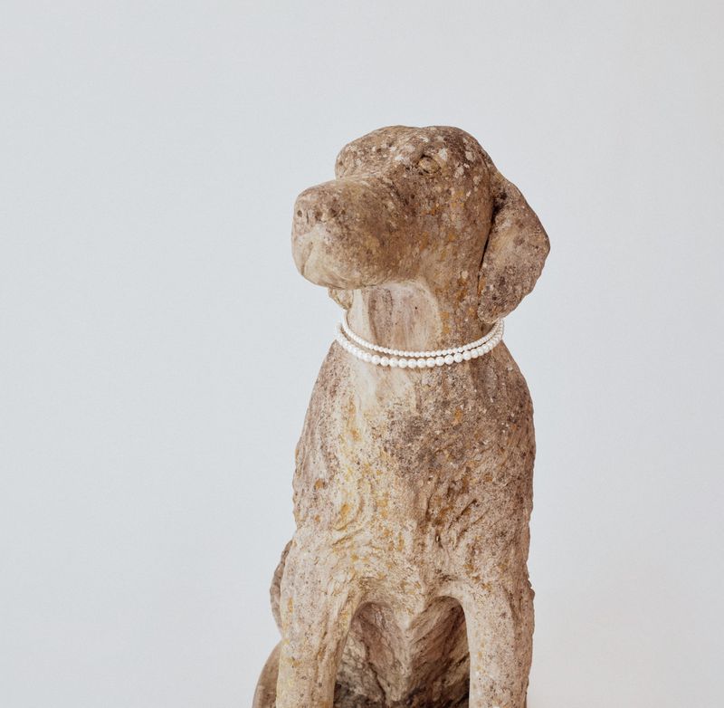 Stone dog sculpture wearing a double strand of white pearl necklaces against a neutral seamless backdrop.