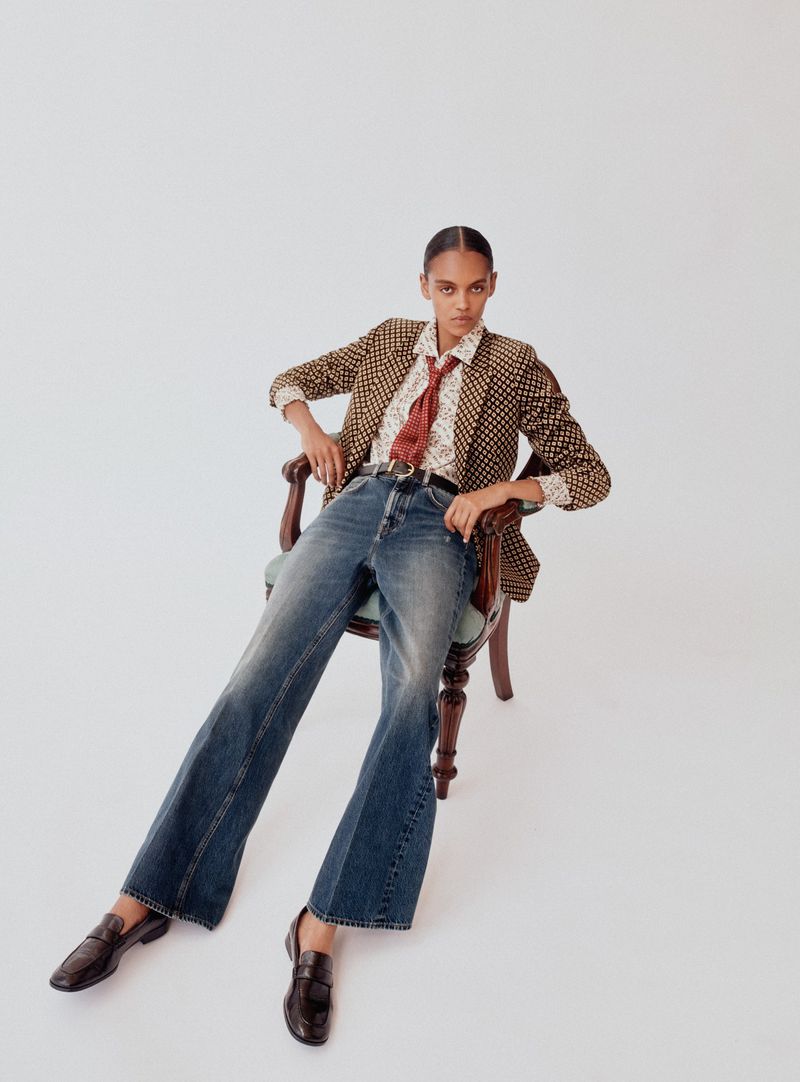 Model wearing patterned blazer, patterned shirt, red tie, and blue jeans seated in an antique wooden chair against white seamless background.