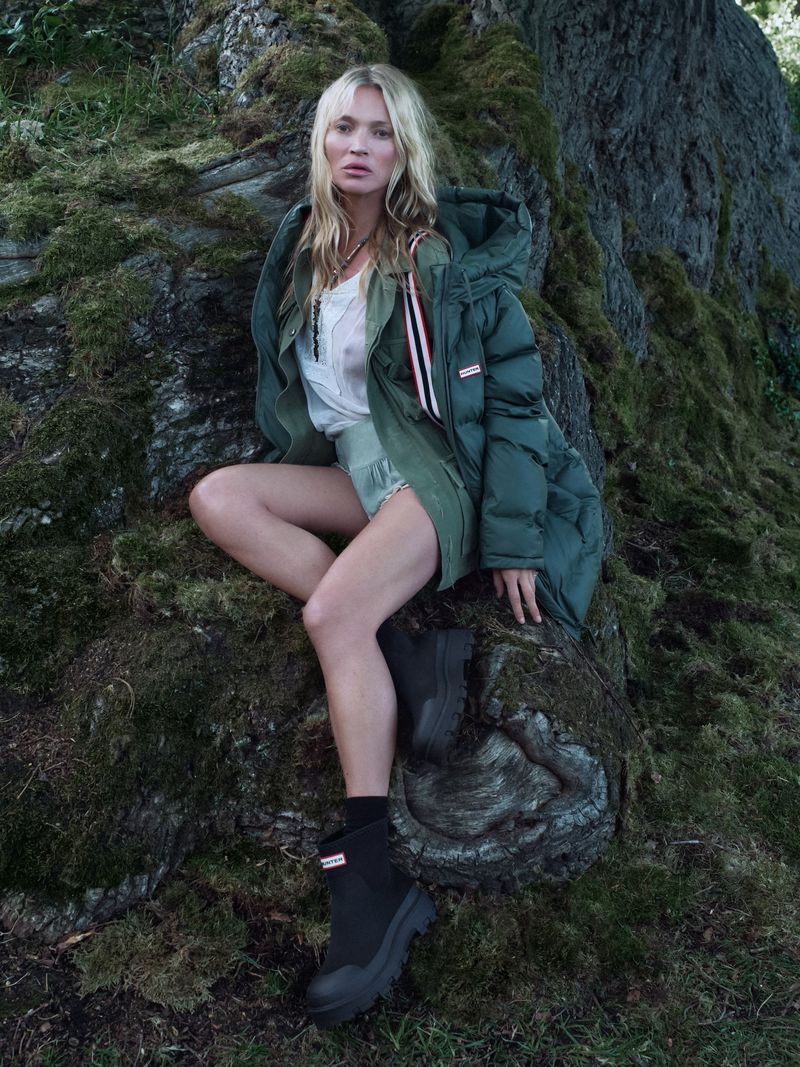 Kate Moss seated outdoors on a mossy rock wearing a green puffer jacket, white top, shorts, and black boots for Hunter Boots.