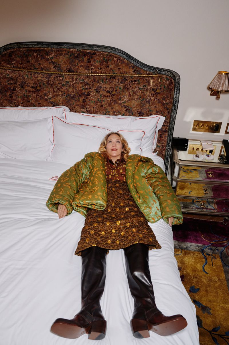 Chloe Sevigny lying on a hotel bed wearing a green padded jacket, patterned dress, and dark leather boots.