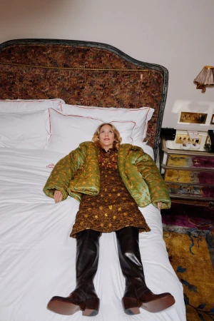 Chloe Sevigny lying on a hotel bed wearing a green padded jacket, patterned dress, and dark leather boots.