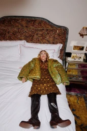 Chloe Sevigny lying on a hotel bed wearing a green padded jacket, patterned dress, and dark leather boots.