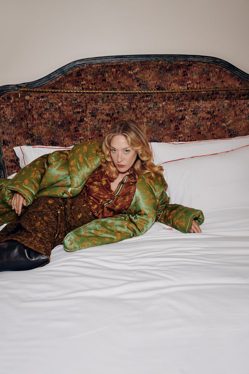 Chloe Sevigny wearing green patterned outerwear and brown trousers lying on a bed with patterned headboard