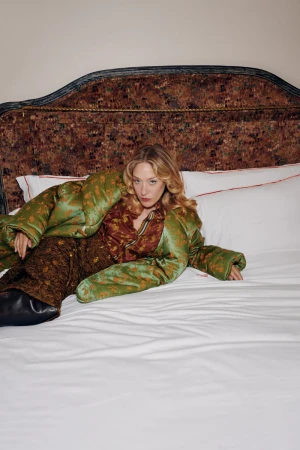 Chloe Sevigny wearing green patterned outerwear and brown trousers lying on a bed with patterned headboard