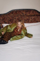 Chloe Sevigny wearing green patterned outerwear and brown trousers lying on a bed with patterned headboard