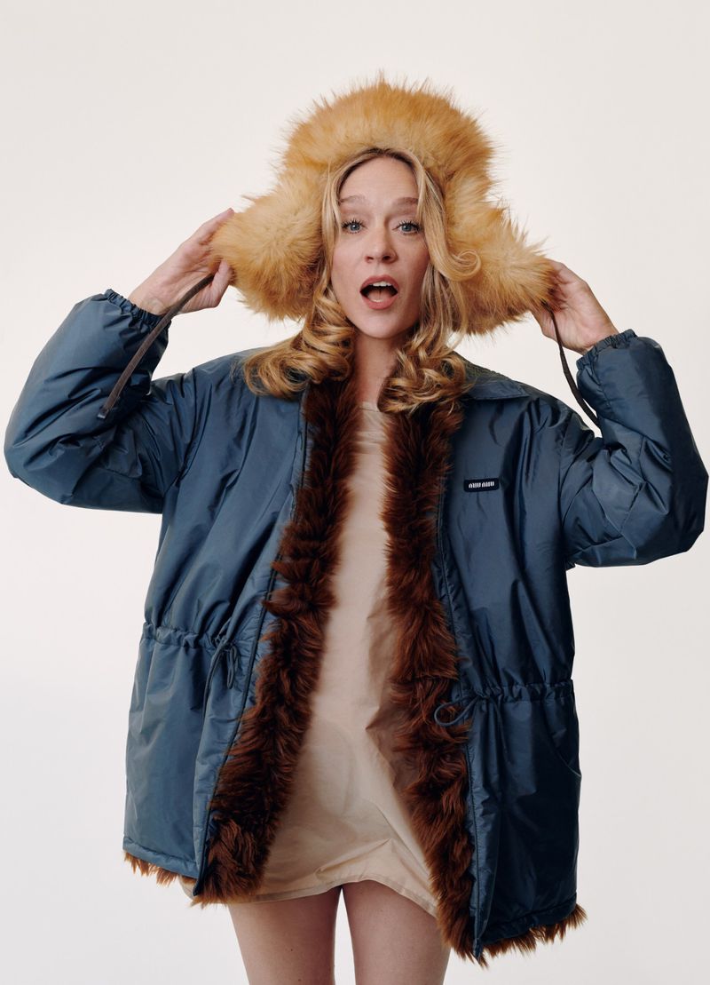 Chloe Sevigny wearing a blue winter coat with fur trim and a fuzzy hat against a plain white background