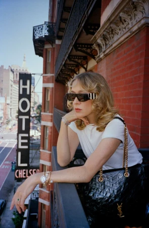 Chloë Sevigny leaning on a balcony railing wearing sunglasses, white t-shirt, and carrying a black chain-strap handbag for mytheresa campaign
