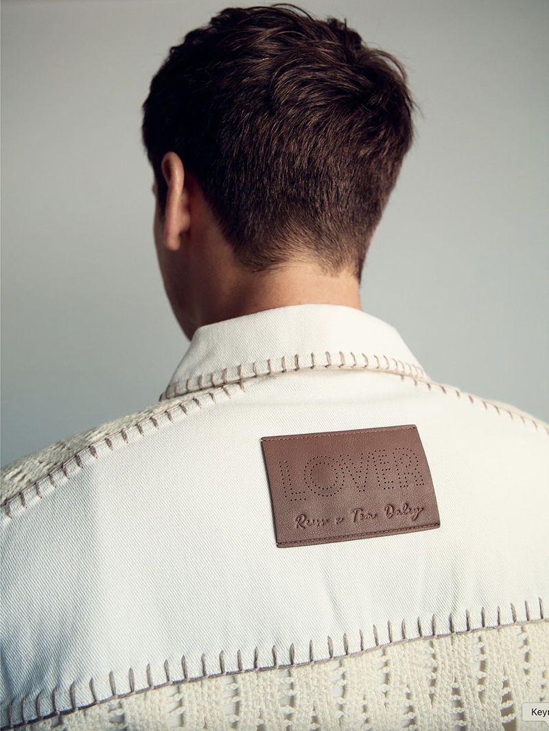 Back view of a person wearing a white jacket with knitted accents and a brown leather label with embossed lettering, on a plain background.