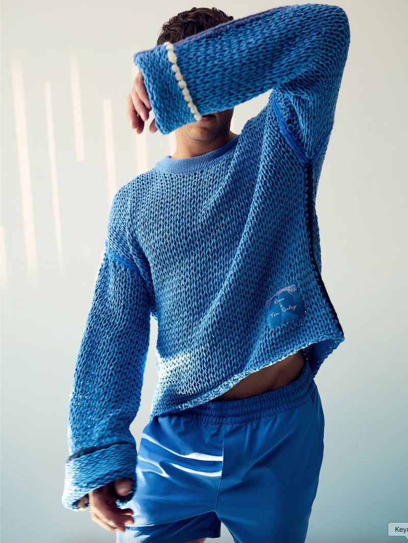 Person with hand covering face wears a blue knit sweater and matching blue shorts in a studio setting with soft, natural light.