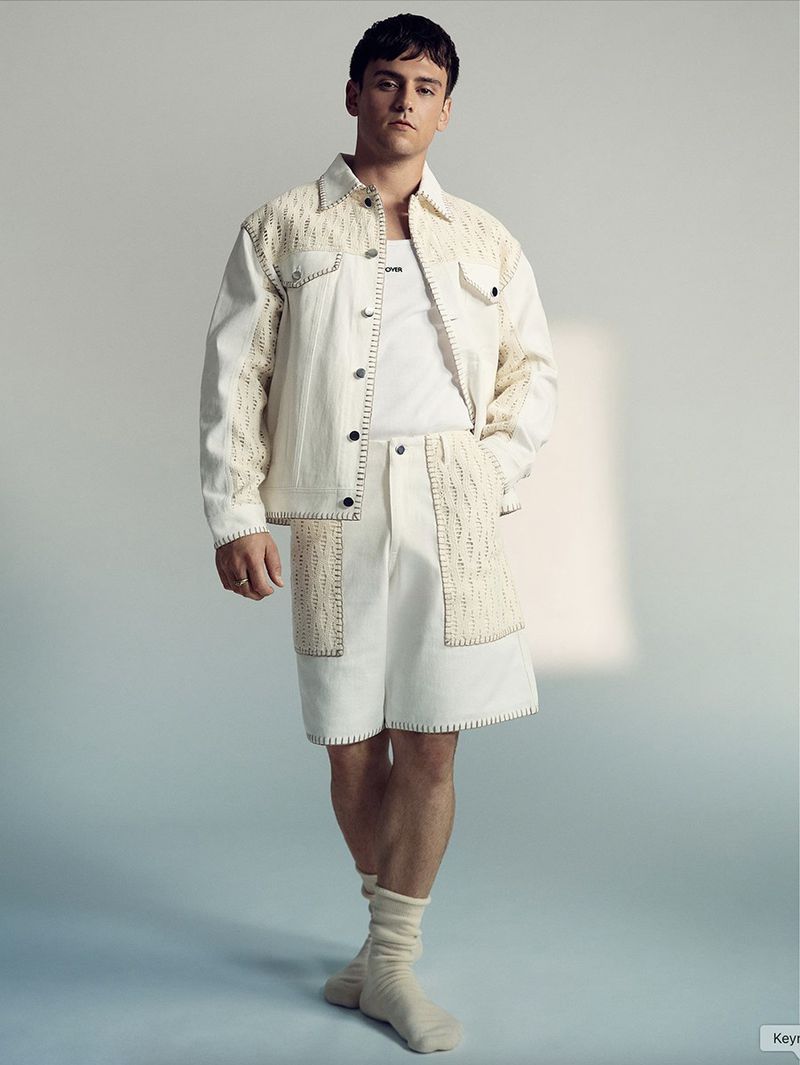 A young adult man stands in a photo studio wearing a matching white denim jacket and shorts with textured details, paired with socks.