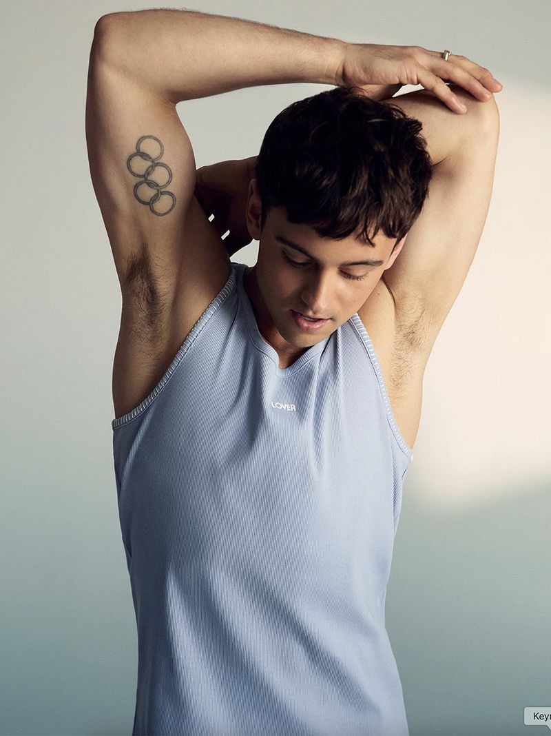 Adult male athlete stretches his arm with Olympic tattoo on his upper arm, wearing a light blue sleeveless shirt against a solid backdrop.