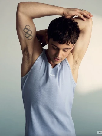 Adult male athlete stretches his arm with Olympic tattoo on his upper arm, wearing a light blue sleeveless shirt against a solid backdrop.