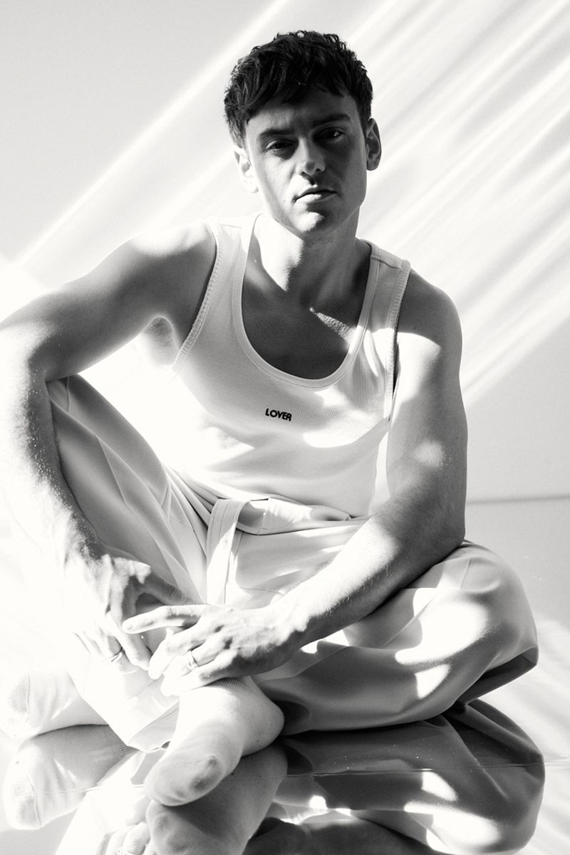 Monochrome portrait of a man in a white tank top sitting cross-legged in bright, diffused lighting against a light background.