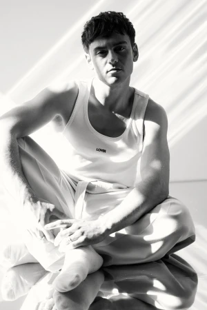Monochrome portrait of a man in a white tank top sitting cross-legged in bright, diffused lighting against a light background.