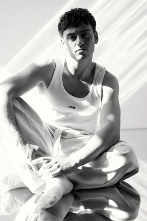 Tom Daley seated on a reflective surface wearing a white tank top and trousers under strong natural light streaming from above