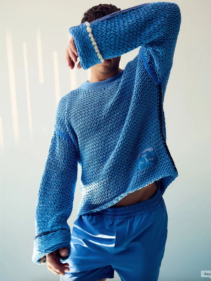 Man wearing a textured blue knit sweater and blue trousers with his arm raised across his face against a pale wall.