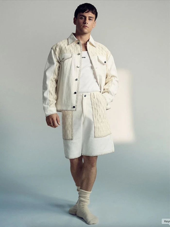 Tom Daley posing in a cream whipstitched jacket and matching shorts with knit panels against a pale studio backdrop.
