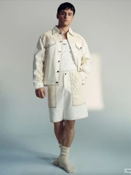 Tom Daley posing in a cream whipstitched jacket and matching shorts with knit panels against a pale studio backdrop.
