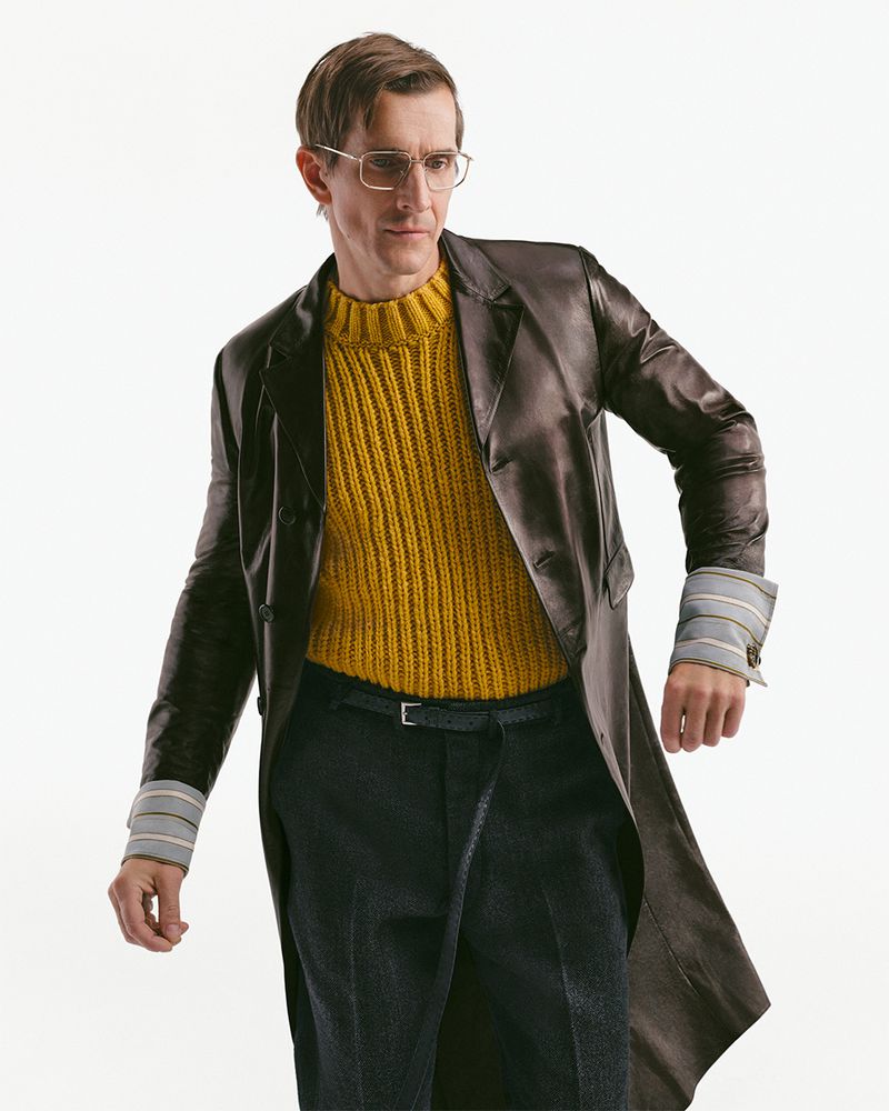 Model wearing a brown leather coat, yellow ribbed sweater, and dark trousers against a white seamless background