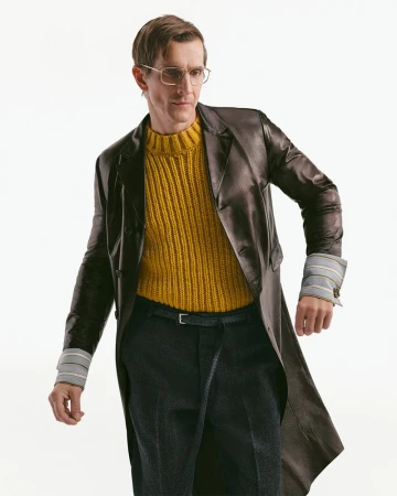 Model wearing a brown leather coat, yellow ribbed sweater, and dark trousers against a white seamless background