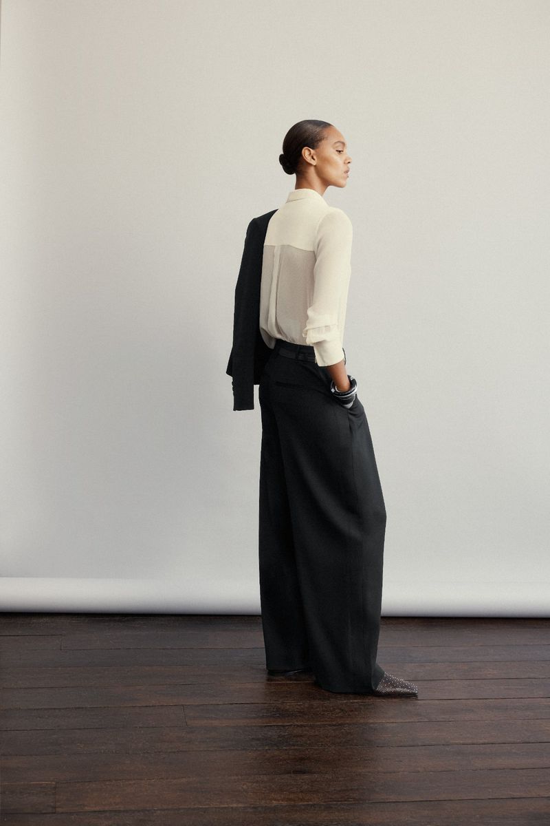 Model wearing a white blouse and black wide-leg trousers holding a jacket against a white studio background
