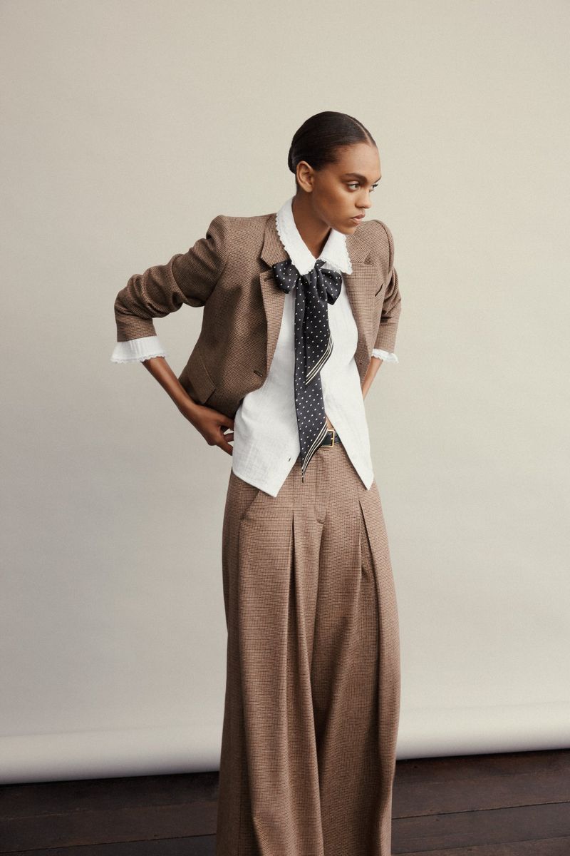 Model wearing a brown tweed blazer and wide-leg trousers with a white blouse and polka dot necktie against a seamless grey backdrop.
