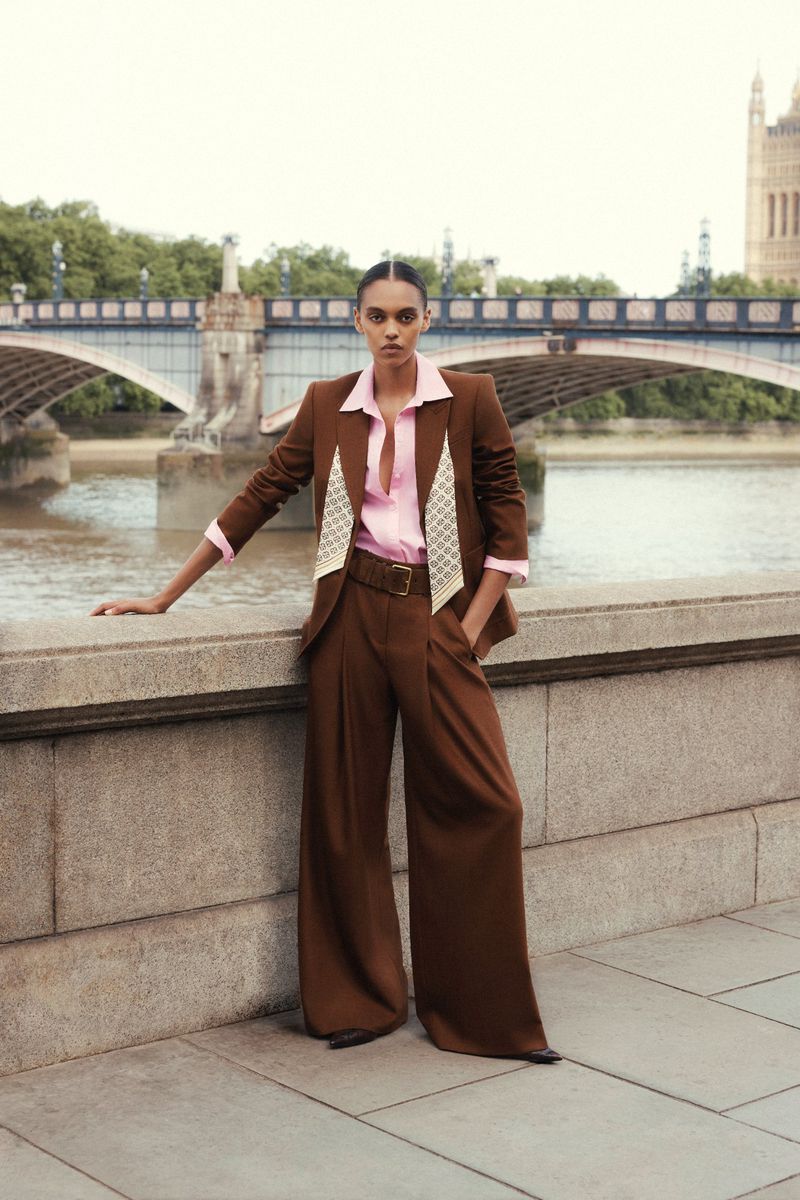 Model in brown trousers and matching blazer with pink shirt posing against stone bridge and river for ME+EM