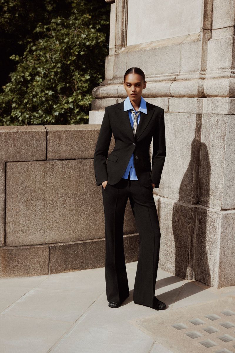 Model standing outdoors wearing a black tailored suit jacket and trousers with a blue shirt and tie