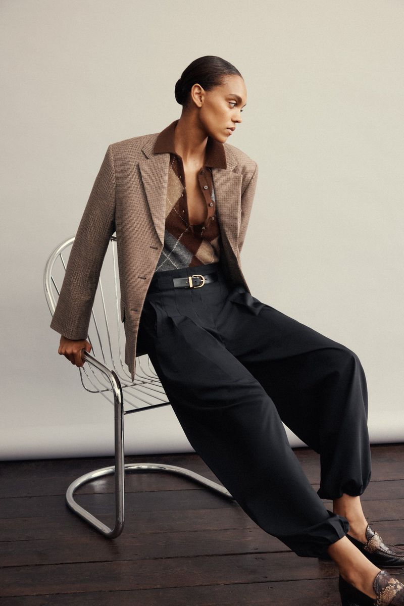 Model wearing a brown blazer, argyle knit top, and dark trousers seated on a chrome wire chair against a light grey studio background
