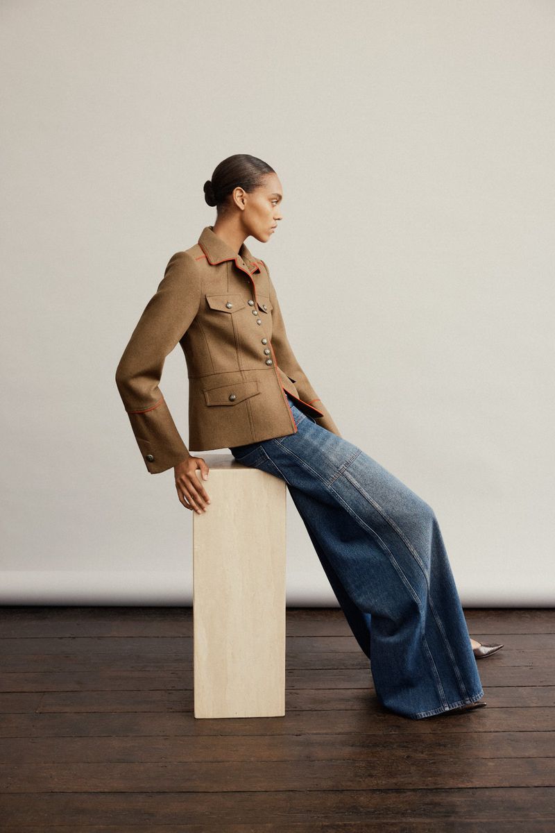 Model seated on a wooden block wearing a brown tailored jacket and wide-leg denim jeans against a seamless backdrop.